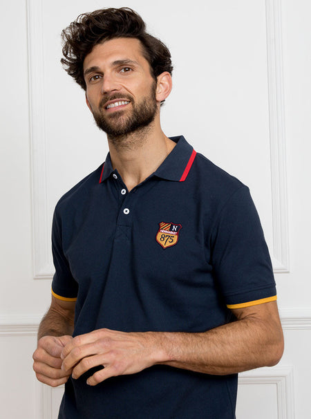Man wearing navy polo shirt in 100% cotton with Newmarket 875 crest embroidered on the front and giant gold number 5 on the back with gold sleeve trimming and red collar trimming.